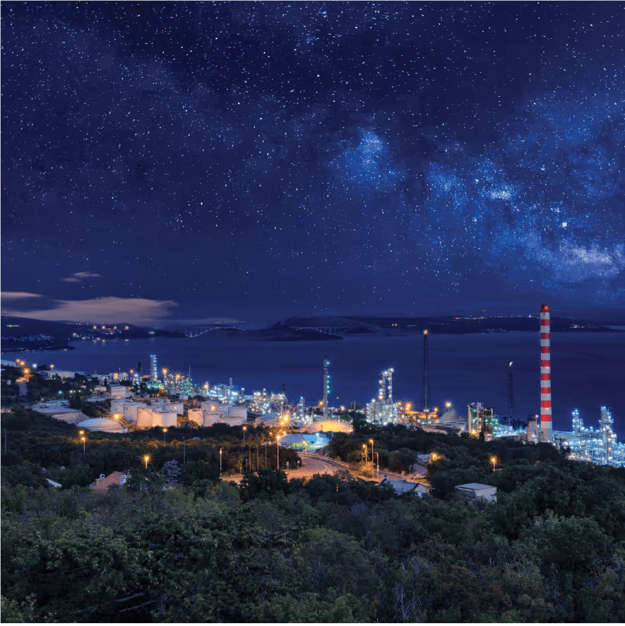 Nighttime view of an illuminated industrial refinery near a body of water under a star-filled sky with the Milky Way visible.