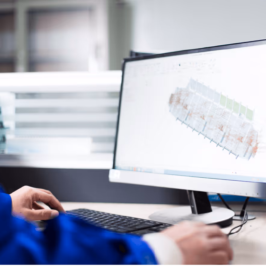 Person wearing blue working on a computer displaying a technical 3D model on the screen.