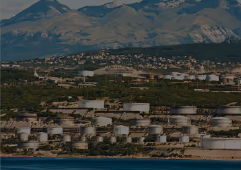 Industrial oil storage tanks near a coastal area with mountains and a town in the background.