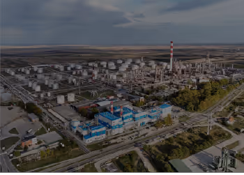 Aerial view of a large industrial complex with numerous storage tanks, pipelines, and a tall smokestack, surrounded by roads and fields.