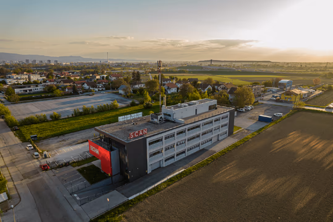 Aerial view of a rectangular commercial building with the word 'SCAN' on its roof and red entrance, surrounded by parking lots, fields, and suburban houses under a partly cloudy sky at sunset.