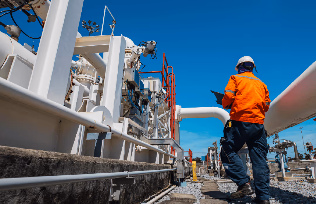 Worker in orange safety jacket and white helmet inspecting equipment at an industrial plant with pipes under a clear blue sky.