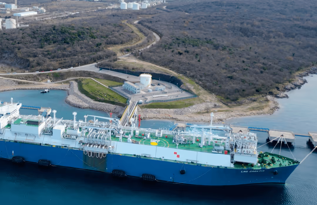 Large LNG cargo ship docked at an industrial port with storage tanks and pipelines onshore.